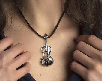 Può includere: Collana con pendente a forma di violino in argento con dettagli intricati e una piccola gemma blu. Il pendente è appeso a una collana di cordino nero. Il design presenta volute e una finitura lucida, con un tema musicale.