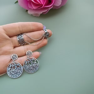 Puede incluir: Un conjunto de pendientes y un anillo de plata con un diseño circular con letras armenias. Los pendientes tienen un pequeño remolino plateado sobre el diseño circular.