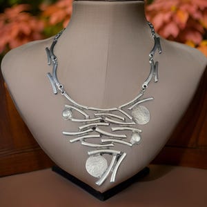 May include: A silver necklace with a geometric design. The necklace is made of interconnected silver bars and circles. The necklace is displayed on a mannequin.