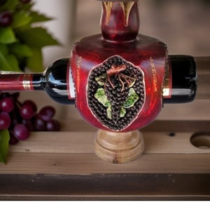 May include: A wooden wine bottle holder shaped like a red pomegranate with a grapevine design. The holder is holding a bottle of red wine with a gold label that says "Armenia".