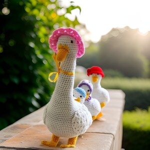 May include: A group of crocheted goose figurines, each with a different colored hat. The largest goose is white with a pink hat and yellow ribbon. The other two geese are smaller and have red and purple hats.