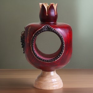 May include: A red wooden pomegranate sculpture with a circular opening and a small wooden base. The pomegranate is carved with details and has a gold inscription.