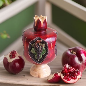 May include: A red, carved wooden pomegranate with gold accents and a grapevine design. The pomegranate is on a wooden base and is surrounded by red pomegranates.