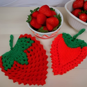Puede incluir: Dos agarraderas de olla de ganchillo con forma de fresa. Una es roja con hojas verdes y la otra es roja con un corazón verde. Ambas están sobre una superficie blanca.
