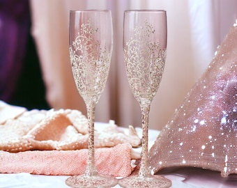 May include: Two clear glass champagne flutes with delicate gold painted designs. The flutes are on a white surface with a pink and white fabric in the background.