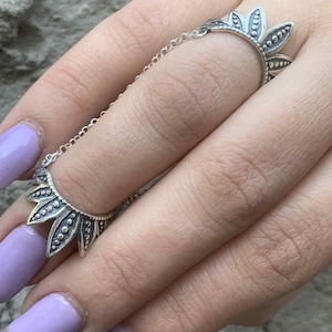 May include: A silver-colored ring with a chain connecting two floral-shaped bands. The ring features a detailed design with small, raised dots and leaf-like shapes. The ring is worn on a finger, showcasing the intricate craftsmanship.