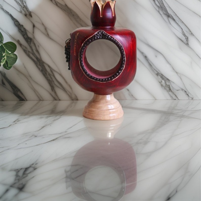 May include: A red wooden pomegranate-shaped decorative object with a gold crown on top. The object has a circular opening in the center and is sitting on a wooden base.