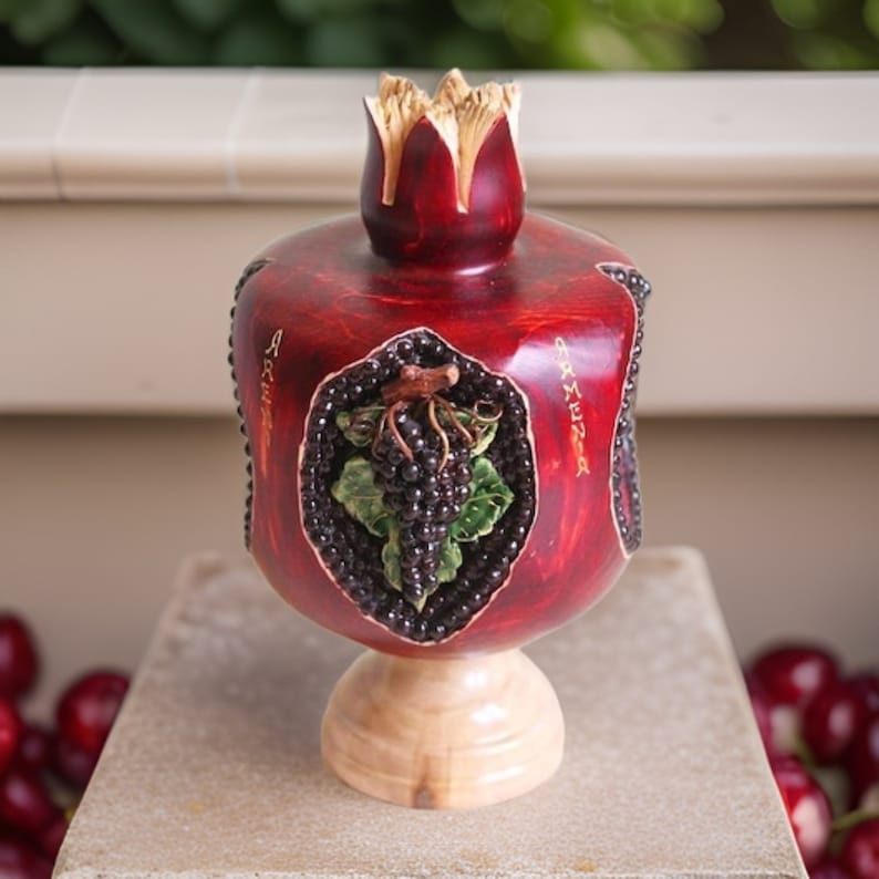 May include: A hand-carved wooden pomegranate with a red finish and a gold inscription that reads "Armenia". The pomegranate is decorated with a cluster of black grapes and green leaves.
