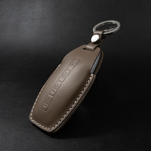 Könnte beinhalten: Eine braune Leder-Schlüsselanhängerhülle mit weißen Nähten und einem silbernen Schlüsselring. Die Hülle hat geprägte Symbole und einen Druckknopfverschluss. Der Schlüsselanhänger dient zum Schutz des Autoschlüssels.