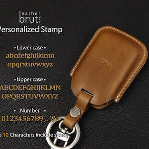 Może przedstawiać: Brązowy sk&oacute;rzany brelok do kluczy ze srebrnym k&oacute;łkiem. Brelok ma białe przeszycia i wygrawerowane słowo "brut" z przodu. Tekst "Personalized Stamp" jest wyświetlany nad brelokiem, wraz z dostępnymi znakami do personalizacji.