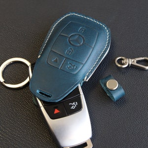 May include: A blue leather key fob with a silver key inside. The key fob has white stitching around the edges and a silver key ring attached to it. The key has four buttons on it.