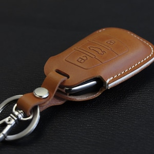 Pu&ograve; includere: Un portachiavi in ​​pelle marrone con dettagli di cuciture bianche e un portachiavi argentato. Il portachiavi ha un'apertura per una chiave dell'auto e un design a pulsante.