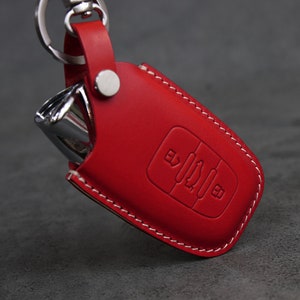 May include: Red leather key fob with white stitching. The fob has a silver metal key and a button with a symbol on it.