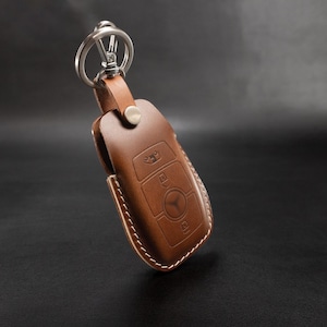 May include: Brown leather key fob with a silver key ring. The fob has a cutout design for a car key with buttons.