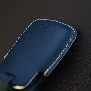 May include: A blue leather key fob with white stitching. The fob has four buttons with symbols for unlock, lock, trunk, and panic.