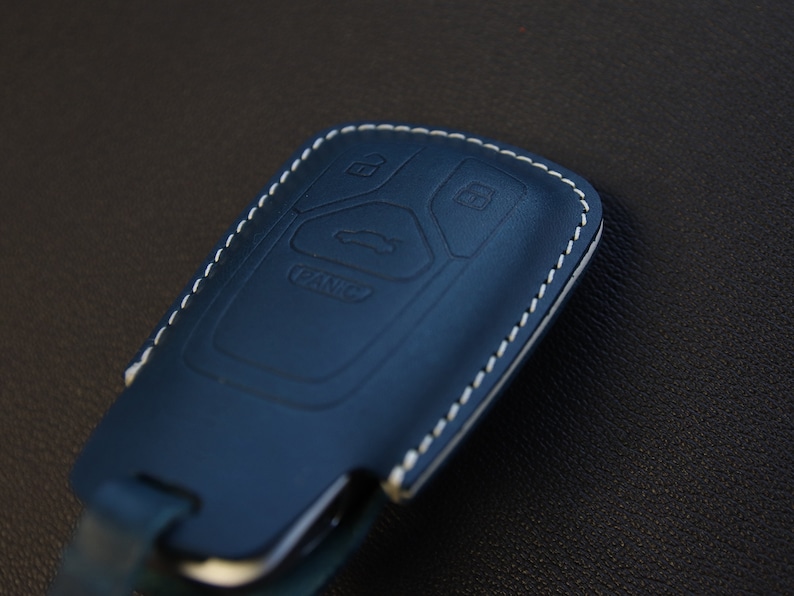 May include: A blue leather key fob with white stitching. The fob has four buttons with symbols for unlock, lock, trunk, and panic.