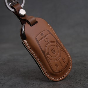 Może przedstawiać: Brązowy skórzany brelok do kluczy ze szwami na krawędziach. Brelok ma wycięcie na kluczyk samochodowy i posiada wytłoczone przyciski.