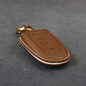 May include: A brown leather key fob with white stitching. The fob has four buttons with symbols for lock, unlock, trunk, and panic.