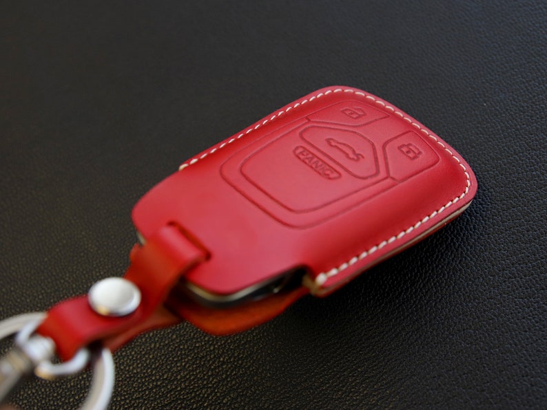 May include: Red leather key fob with white stitching and a key ring. The fob has a car logo and buttons.