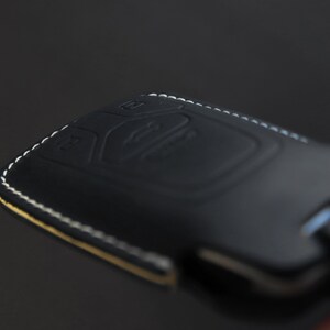 May include: Black leather key fob with white stitching. The fob has a textured surface and a button on the top.