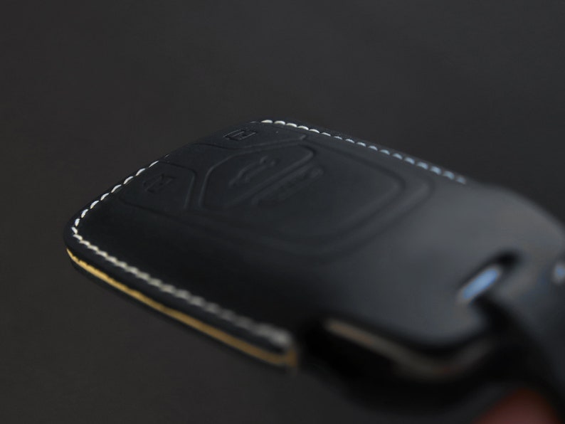 May include: Black leather key fob with white stitching. The fob has a textured surface and a button on the top.