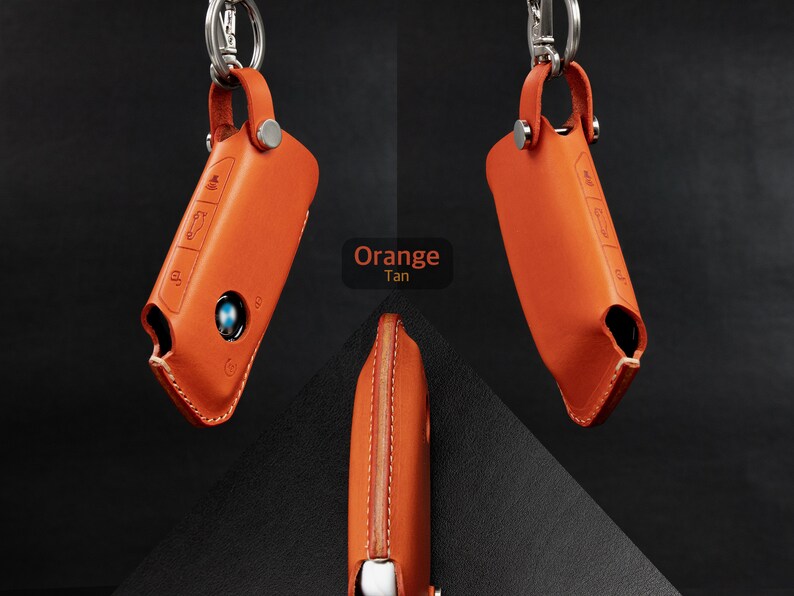 May include: Orange leather key fob with a silver key ring and a BMW logo. The key fob has a stitched seam and a button on the side.