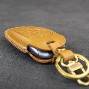 May include: A tan leather key fob with a white stitching detail and a gold metal key ring. The key fob has a cutout for a car key and a small button on the side.