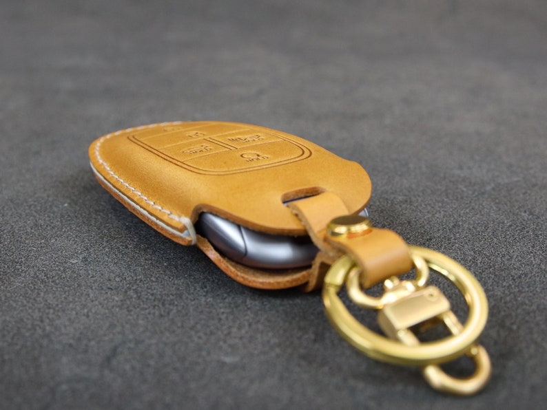 May include: A tan leather key fob with a white stitching detail and a gold metal key ring. The key fob has a cutout for a car key and a small button on the side.