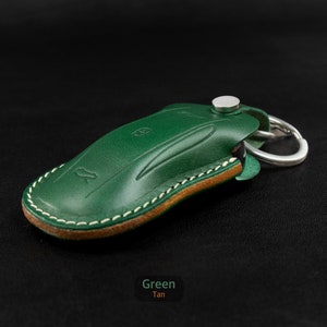 May include: A green leather key fob with a tan leather trim and a silver metal ring. The fob has a stitched edge and a small silver metal button.