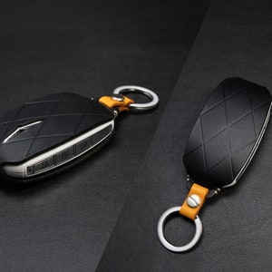 May include: Black key fob with a diamond pattern and silver accents. The fob is attached to a silver keyring with a tan leather strap. The key fob has buttons and a silver emblem. The background is black.