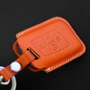 May include: Orange leather key fob with a white stitching detail. The fob has a key chain attached and features a design with four buttons.