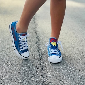 May include: Blue canvas sneakers with white laces and a white rubber toe cap. The sneakers have a red and yellow graphic on the tongue and the word "JAYHAWKS" in red lettering on the side. The shoes are on an asphalt surface.