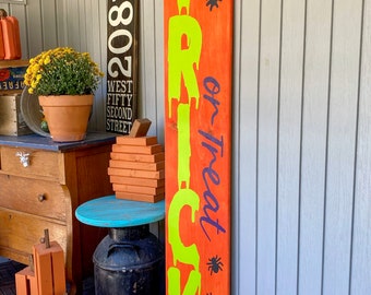 May include: A wooden sign with the words "Trick or Treat" painted in green and purple on an orange background. There are three black spiders painted on the sign.