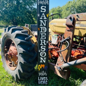May include: Black wooden sign with the text "The fourth SANDERSON sister lives here" in white and yellow lettering. The sign is decorated with black spiders, spiderwebs, and orange hearts. The sign is leaning against a red tractor.