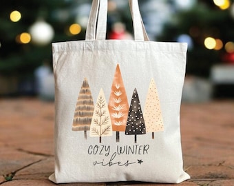 May include: A cream-colored tote bag featuring a design of stylized Christmas trees in shades of brown, orange, and black. The words "cozy winter vibes" are printed below the trees. The bag has a simple handle and is set against a blurred background of Christmas trees.