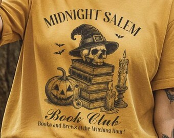 Puede incluir: Camiseta de color mostaza con el texto "MIDNIGHT SALEM Book Club". El gráfico presenta una calavera con un sombrero de bruja, una pila de libros, una calabaza y velas. La camiseta también tiene el texto "Books and Brews at the Witching Hour!"