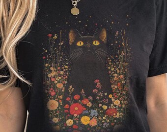 Può includere: T-shirt nera con una grafica di un gatto nero con occhi gialli circondato da fiori colorati. Il gatto è centrato sulla maglietta, con i fiori che lo circondano. La parola "Black" è stampata in bianco in basso a destra.