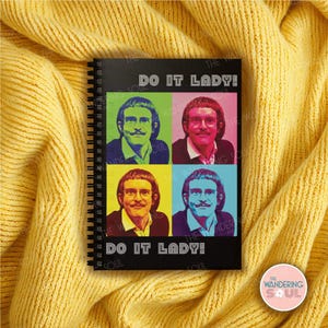 May include: Black spiral-bound notebook featuring a pop art style image of a man with a mustache in four different color blocks. The text "DO IT LADY!" is printed above and below the image. The notebook is resting on a yellow knitted surface.