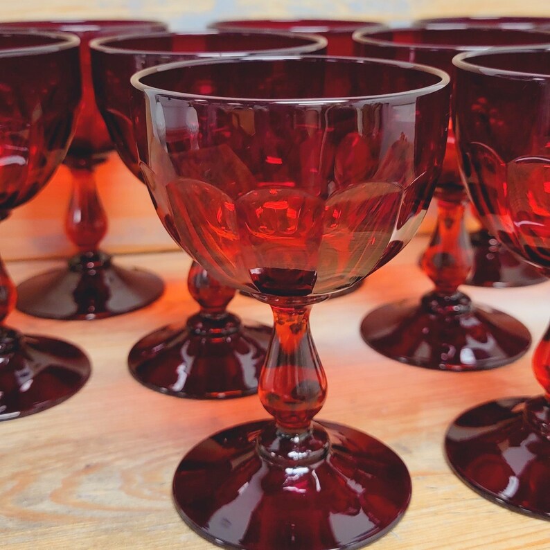 May include: A collection of ruby red wine glasses. Each glass features a round base, a slender stem, and a bowl with a decorative pattern. The glasses are arranged on a light-colored wooden surface, creating a visually appealing display.
