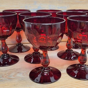 May include: A collection of ruby red wine glasses. Each glass features a round bowl, a stem with a bulbous design, and a circular base. The glasses are arranged on a light-colored wooden surface, showcasing their rich color and intricate details.