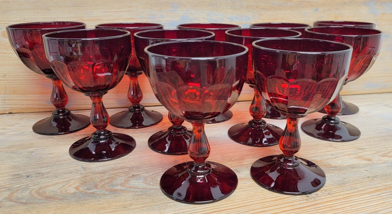 May include: A collection of ruby red wine glasses. Each glass features a round bowl, a stem with a bulbous design, and a circular base. The glasses are arranged on a light-colored wooden surface, showcasing their rich color and intricate details.