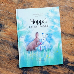 May include: A blue book cover with a watercolor illustration of two bunnies holding an Easter egg. The title of the book is "Hoppel und der Osterhase" by Kathrin Siegenthaler and Marcus Pfister. The book is published by Nord-Süd Verlag.