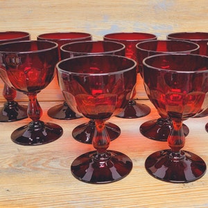 May include: A collection of ruby red stemmed glasses. The glasses have a faceted design on the bowl and a decorative stem. The glasses are arranged on a light wood surface.