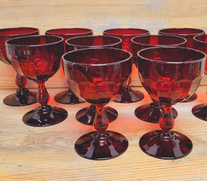 May include: A collection of ruby red stemmed glasses. The glasses have a faceted design on the bowl and a decorative stem. The glasses are arranged on a light wood surface.