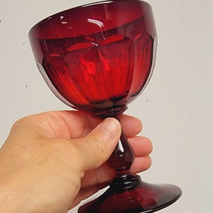 May include: A ruby red wine glass with a faceted bowl and a stem. The glass is held in a hand against a white background. The glass has a round base and a bulbous stem.