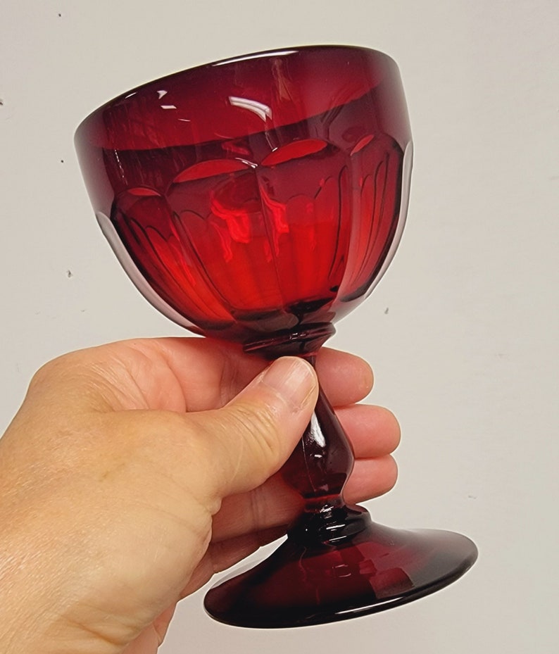 May include: A ruby red wine glass with a faceted bowl and a stem. The glass is held in a hand against a white background. The glass has a round base and a bulbous stem.