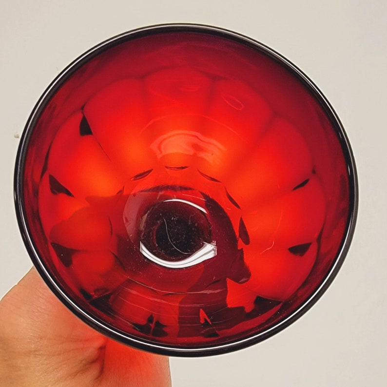 May include: A top-down view of a ruby red glass with a black rim. The glass has a textured interior and a clear, circular base. The glass is held by a hand, with the background out of focus.