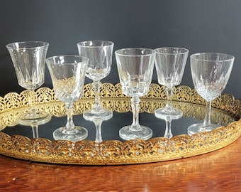 May include: Set of six clear glass wine glasses on a gold metal tray with a decorative design. The glasses are all different shapes and sizes.