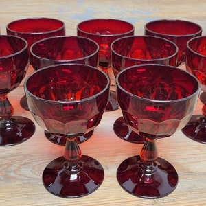 May include: A collection of ruby red stemmed glasses. Each glass features a round base, a decorative stem, and a faceted bowl. The glasses are arranged on a light-colored wooden surface, showcasing their rich color and design.