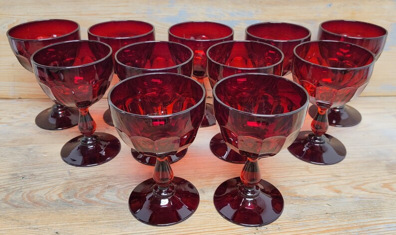 May include: A collection of ruby red stemmed glasses. Each glass features a round base, a decorative stem, and a faceted bowl. The glasses are arranged on a light-colored wooden surface, showcasing their rich color and design.